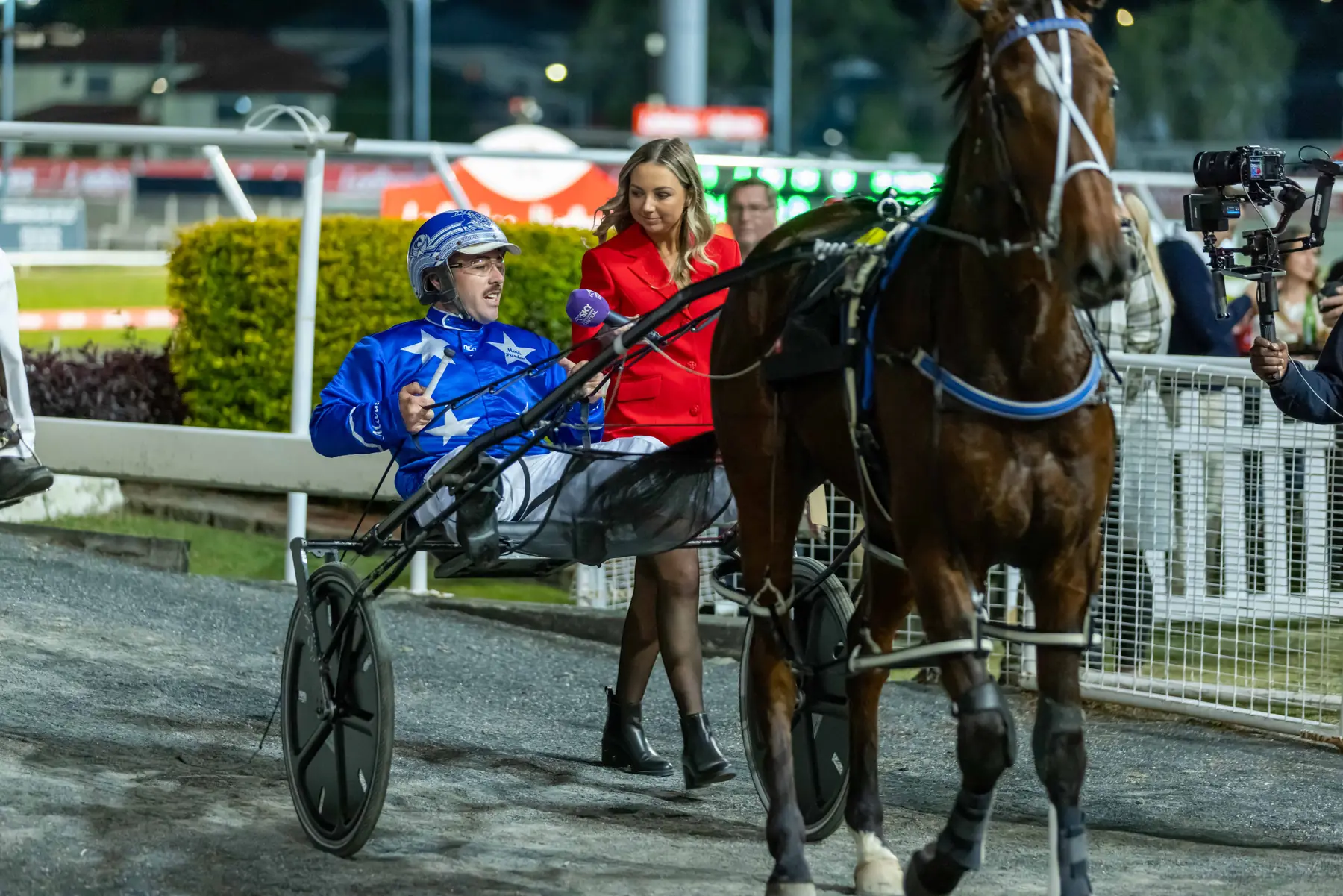 Harness racing driver being interviewed beside the track while a video camera records the moment